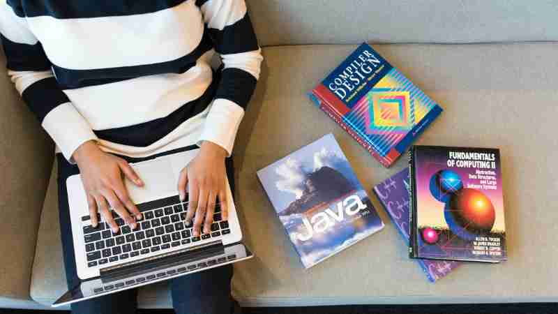 Woman using laptop on sofa, surrounded by programming books, learning coding., tags: und - pexel